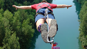 man in red shirt with arms out leaping off a bridge in a bungee jumping harness