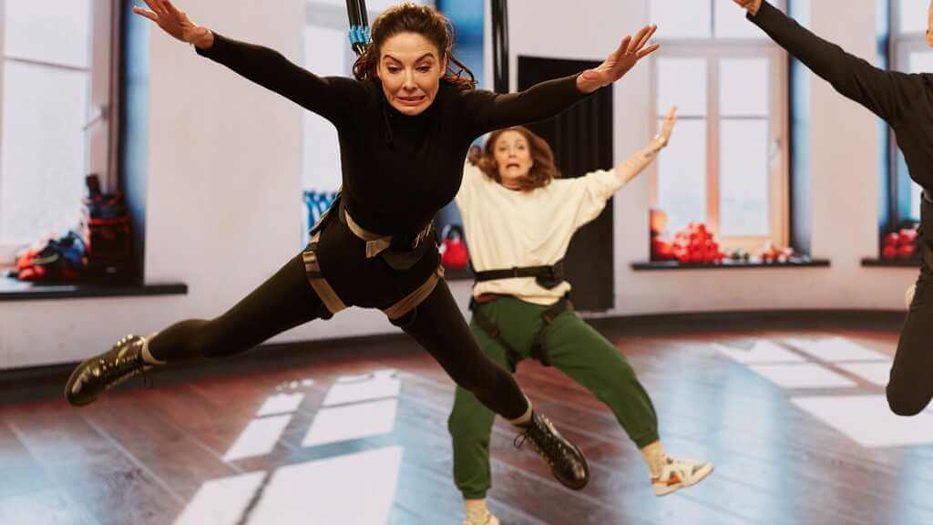 whitney cummings and drew barrymore suspended in bungee harnesses in a goofy way with legs and arms splayed